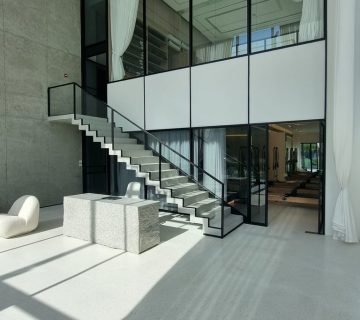 Modern interior design featuring a spacious entryway with a minimalist staircase, large glass windows, and concrete walls A stylish stone table is positioned on a polished floor, illuminated by natural light filtering through sheer curtains