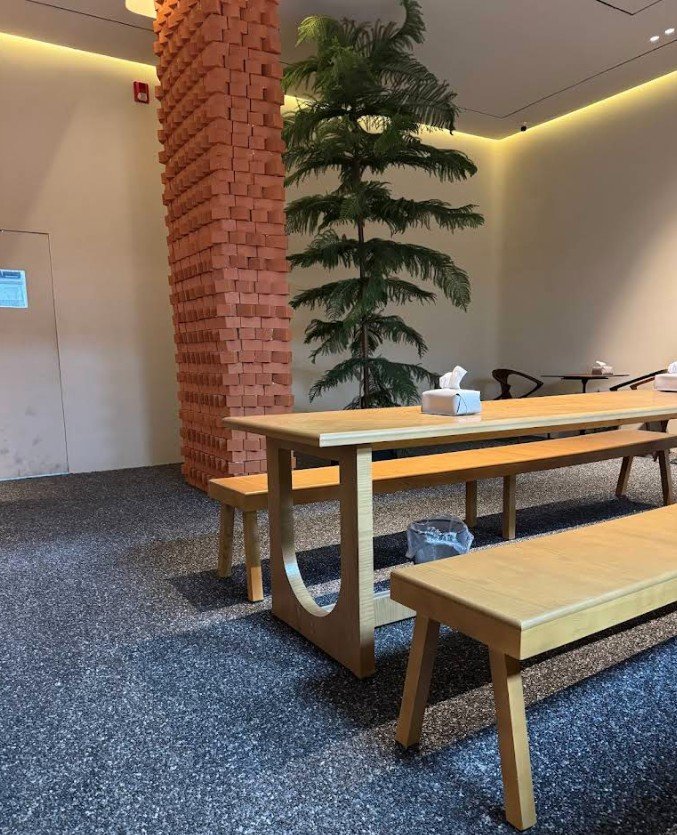 Modern interior design featuring a wooden dining table and benches, complemented by a tall artificial palm tree and a textured brick column  The space has a minimalist aesthetic with soft lighting and a polished floor 