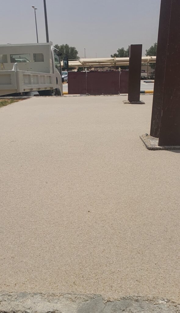 Image of a freshly paved concrete surface with a light texture, featuring a white truck parked nearby and two vertical metal structures in the foreground  The background shows a construction area with barriers and trees under a clear sky 