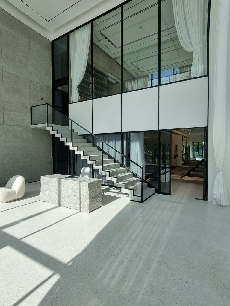 Modern interior design featuring a spacious entryway with a minimalist staircase, large glass windows, and natural light illuminating polished concrete floors  A stylish stone table and contemporary seating enhance the elegant atmosphere 