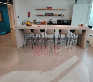 Modern kitchen with a large concrete island and six stylish bar stools, featuring a wall shelf stocked with snacks and kitchen appliances in a bright, open layout 